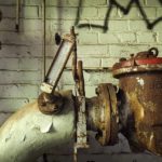 Old pumps and pipes with an attached meter in need of water well repair in front of a brick wall with graffiti