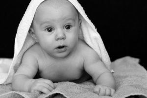 A baby holding its head up during tummy time laying on a towel with another towel draped over its head and back.