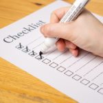 A hand holding a marker making check marks on a paper checklist on a wooden table.